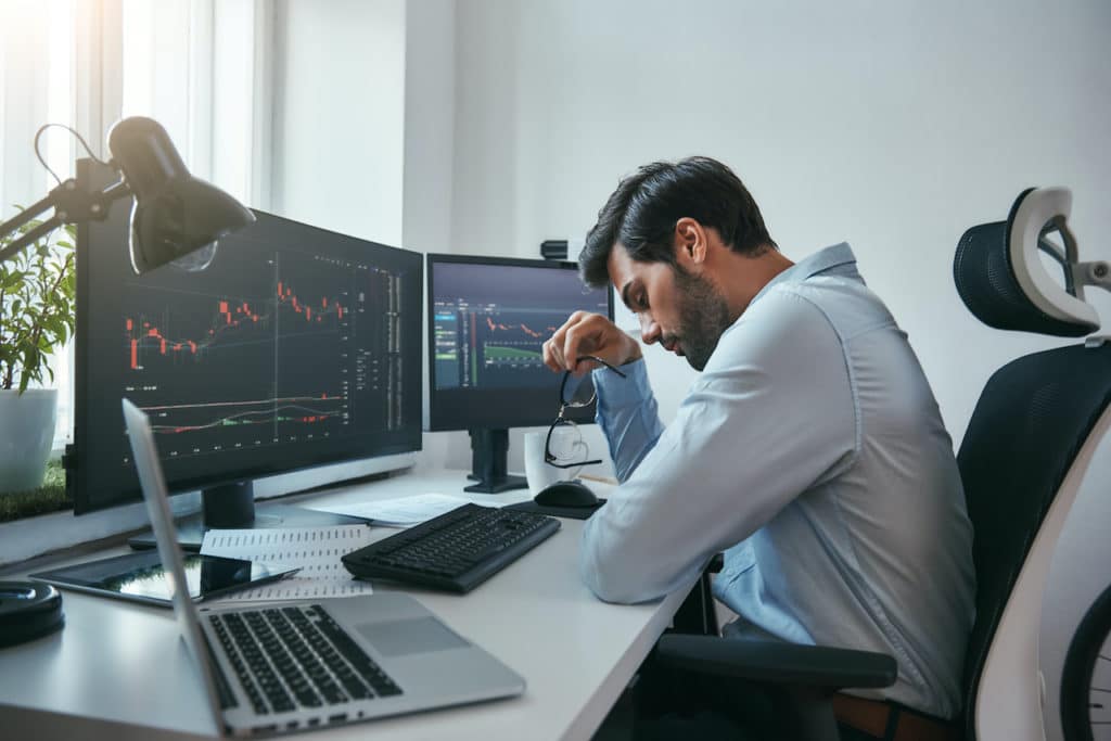 man sits in front of two computer screens showing graphs of debts and assets as he contemplates bankruptcy due to covid-19