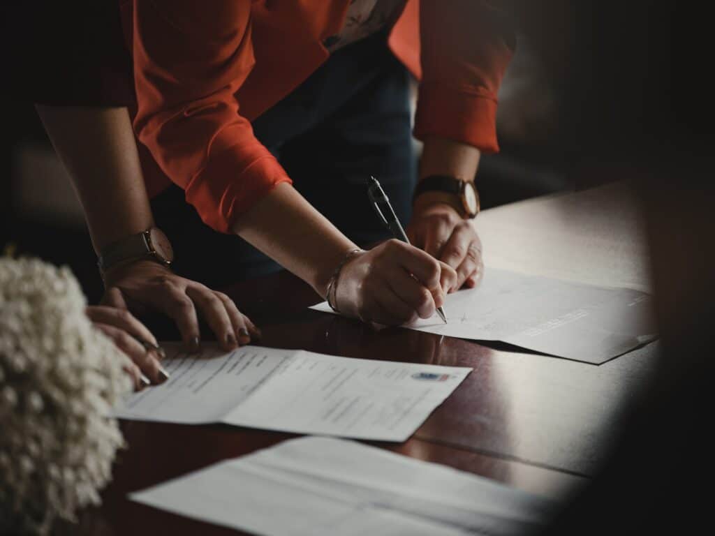 Two people lean over a table and sign paperwork. It's important to know what you're signing to avoid breach of contract.
